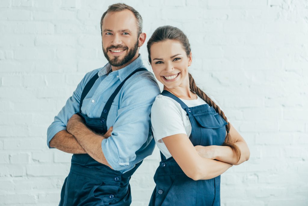 Happy couple in blue overalls smiles together in front of a white brick wall, showcasing their joyful connection.